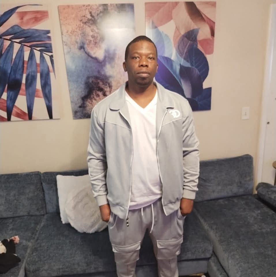 Portrait of Jamel Miller standing in his living room in a light gray tracksuit
